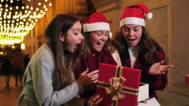 Noel şapkası takan heyecanlı genç kadınlar, büyülü bir Noel kutlaması sırasında etrafı ışıldayan tatil ışıklarıyla çevrili, neşeli ve sürprizlerle dolu bir hediye kutusunu gece birlikte açıyorlar. Yüksek