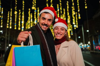 Noel Baba şapkası takan ve alışveriş torbaları tutan mutlu genç çift tatil ışıklarının altında Noel alışverişinin tadını çıkarıyor. Tatil sezonunu bir şehir gecesinde neşe ve beraberlikle kutluyorlar.