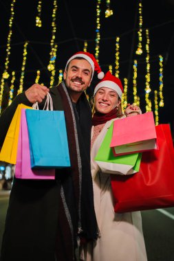 Dikey. Noel Baba şapkası takan ve alışveriş torbaları tutan mutlu genç çift bayram ışıklarının altında Noel alışverişinin tadını çıkarıyor, bayram sezonunu neşe ve beraberlikle kutluyorlar.