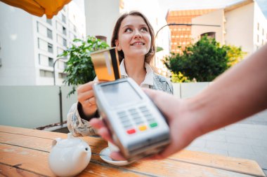 Bir barın terasında oturmuş gülümseyen genç bir kadın, bir kredi kartıyla çayının parasını mutlu bir şekilde ödüyor, modern ödeme teknolojisini gözler önüne seriyor ve canlı bir şehir ortamında gündelik yemek yiyor. Yüksek kalite