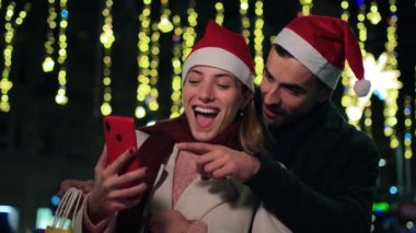 Neşeli genç çift Noel Baba şapkaları takıyor, ellerinde renkli alışveriş çantaları tutuyorlar ve neşeli bir Noel gezisi sırasında parlak tatil ışıkları altında birlikte akıllı telefon kullanıyorlar, sevgi gösterisi yapıyorlar. Yüksek