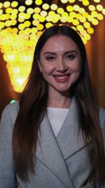 Dikey. Uzun saçlı gülümseyen kadın doğrudan kameraya bakıyor, şık bir ceket giyiyor, açık havada duruyor, akşam şehir ortamında mutluluk, güven ve sıcaklığı ifade ediyor. Yüksek kalite