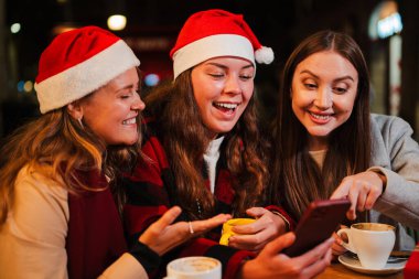 Üç yetişkin kadın gülümsüyor ve cep telefonuyla internete girip kahve dükkanının terasında Noel şapkası takarak eğleniyorlar. Bir grup bayan arkadaş akıllı telefonu izliyor.