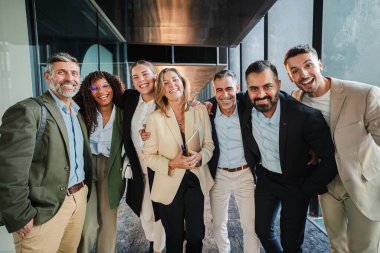 Ofis koridorunda takım çalışmasını, başarıyı, şirket birliğini ve işbirliğini kutlarken aynı anda mutlu bir şekilde poz veren çeşitli gülümseyen iş adamları grubu.