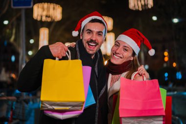 Noel Baba şapkası takan neşeli genç bir çift Noel 'i gece açık havada kutluyor bayram ruhunun keyfini çıkarırken kameraya tatil alışverişlerini gösteriyor. Yüksek
