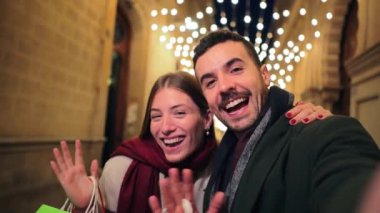 Noel 'i gece açık havada kutlayan alışveriş torbaları taşıyan neşeli genç çift kameraya tatil alışverişlerini gösterirken mevsimlik satışların ve neşeli tatil ruhunun tadını çıkarıyor.