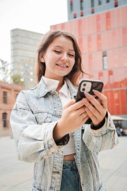 Dikey. Genç kız dışarıda cep telefonu kullanırken gülümsüyor, kot ceket giyiyor, kırmızı binaların önünde dikiliyor ve akıllı telefonuyla konuşurken gündelik bir anın tadını çıkarıyor. Yüksek