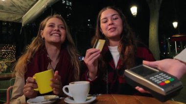 Kahve dükkanının faturasını gece vakti kredi kartıyla öderken gülümseyen iki genç kadın, açık havada ahşap bir masada oturmuş sıcak içeceklerle ve şehir ışıkları altında gecelerinin tadını çıkarıyorlar. Yüksek kalite 4k