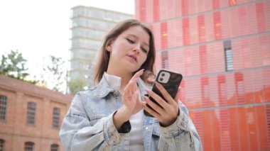 Genç kız dışarıda cep telefonu kullanırken gülümsüyor, kot ceket giyiyor, kırmızı binaların önünde dikiliyor ve akıllı telefonuyla konuşurken gündelik bir anın tadını çıkarıyor. Yüksek kalite 4k