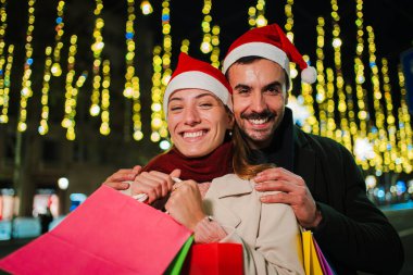 Noel gecesi Noel Baba şapkası takan gülümseyen çift Noel Baba 'nın alışveriş torbalarını tutuyor, ışıklarla bayram ruhunun tadını çıkarıyor, mutluluk ve heyecan göstererek bu sezonu kutluyorlar.