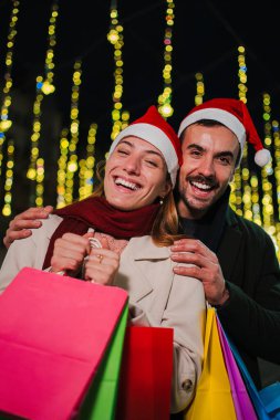 Noel Baba şapkası takan, Noel şenlikli Noel gecelerinde alışveriş torbaları tutan, ışıklarla bayram ruhunun keyfini çıkaran dikey gülümseyen çift. Verme mevsimini kutlarken mutluluk ve heyecan
