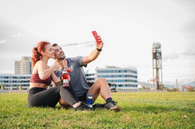 Sportif çiftler çimenlerin üzerinde oturup egzersiz yaptıktan sonra cep telefonlarıyla selfie çekiyorlar, dışarıda spor salonunun keyfini çıkarıyorlar, su şişelerini tutuyorlar, ve mutlu bir şekilde gülümsüyorlar, aktif yaşam tarzını, sağlığını kutluyorlar. Yüksek