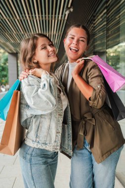 Dikey. Renkli alışveriş torbaları tutan iki genç kadın kameraya gülümsüyor ve özgüvenle bakıyor. Moda, modern bir açık hava perakende ortamında eğlenceli bir gün ve arkadaşlığın tadını çıkarıyorlar.