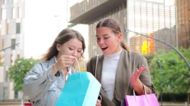 Renkli alışveriş torbaları tutan iki genç kadın, gülümseyen ve özgüvenli bir şekilde içeriye bakan, moda, modern bir açık hava perakende ortamında eğlenceli bir gün ve arkadaşlığın tadını çıkaran. Yüksek kalite 4k
