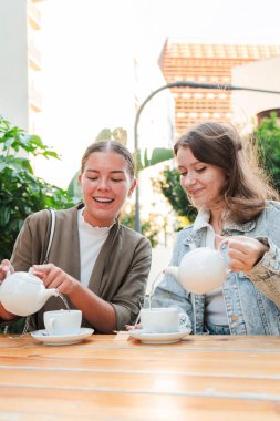 Dikey. Dışarıda çaydan zevk alan iki genç kadın, gülerek ve kaynaşırken, beyaz çaydanlıklardan çay dökerek, sabah huzurlu ve neşeli bir anı paylaşıyorlar. Yüksek kalite fotoğraf