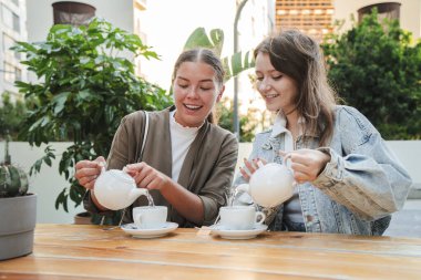Dışarıda çaydan zevk alan iki genç kadın, gülerek ve kaynaşırken, yeşilliklerle çevrili ahşap bir masaya beyaz çaydanlıklardan çay dökerek, sabah güneşinde huzurlu ve neşeli bir anı paylaşıyorlar.