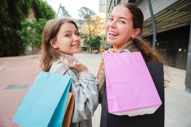 İki neşeli kızın renkli alışveriş çantalarını tutarken, kameraya bakarken, açık hava satış gününün tadını çıkarırken, neşeli ifadelerini sergilerken. Genç kadınlar alışverişleriyle yürüyorlar.