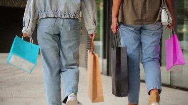 Ağır çekim. Arka planda renkli alışveriş torbalarıyla yürüyen iki kızın bacakları var. Şehir merkezinde şık ve canlı bir tüketim deneyimi sergiliyorlar. Yüksek kalite 4k görüntü