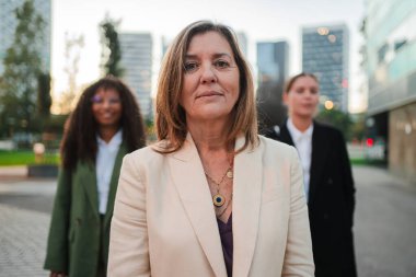 Kendine güvenen bayan lider arka planda meslektaşlarıyla dışarıda duruyor, takım çalışmasını, liderliği ve mesleki başarıyı sembolize ediyor. Olgun bir iş kadını kameraya bakıyor. Yüksek kalite