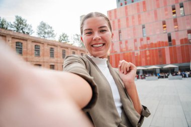 Genç yetişkin bir kadın cep telefonuyla selfie çekerken gülümsüyor, neşeli kişiliğini gösteriyor. Dışarıda neşeli bir ifadeyle kameraya bakıp gülen genç bir kızın yakın portresi.