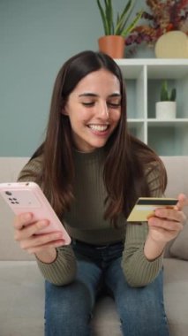 Dikey. Genç bir kadın akıllı telefonunu online alışveriş için kullanıyor, bir kredi kartı tutuyor ve bir kanepede otururken gülümsüyor, rahat ve güvenli bir ödeme deneyimi yaşıyor.