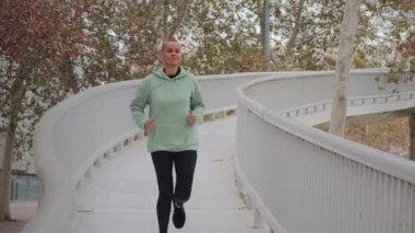 Park köprüsünde koşan aktif bir kadın egzersiz ve açık hava aktiviteleriyle sağlıklı yaşam tarzını sürdürürken azim ve kondisyon göstererek refahı ve kişisel gelişimi vurguluyor.