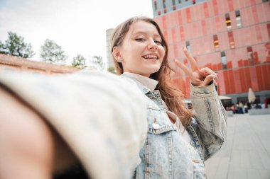 Neşeli Genç Kadın Barış İşareti ile Selfie Çekiyor, Mutluluk ve İyimserlik Anı Yakalıyor. Gülümseyen Genç Kız, Özgüven ve Enerjiyle Hayattan Zevk Alırken Bir Portre Çekiyor. Yüksek