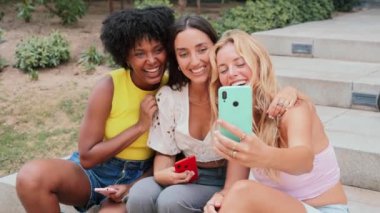 Bir cep telefonuyla eğlenceli bir selfie çeken neşeli üç kadın grubu, Parlak bir açık hava ortamında mutluluk ve arkadaşlık yayıyor, Gülüşmeler ve heyecanla dolu hatırlanmaya değer bir anı yakalıyorlar.