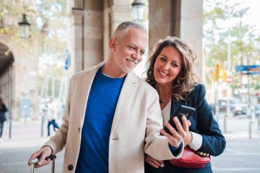 Mutlu çift, akıllı telefonlarını gözlemlerken iç açıcı bir etkileşime giriyorlar, Neşe ve şefkat saçan, Doğal Işığın Gülümsemelerini ve Bağlantılarını Artırdığı Enfes bir açık hava sahnesi