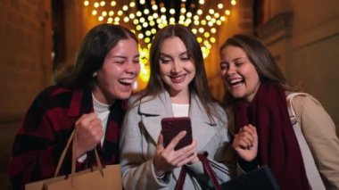Neşeli Bayan Arkadaşlar, birlikte alışveriş yaparken heyecanlı anları paylaşıyor, güzel ışıklarla akıllı telefonlarındaki kahkaha ve mutluluğu yakalıyorlar. Dışarıda cep telefonu kullanan genç bayanlar.