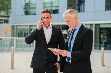 İki iş adamı aktif olarak önemli sohbetlere daldı. Biri telefonda konuşurken, diğeri tabletteki verileri gözden geçiriyor. Profesyonel İşbirliği ve İletişim Özü