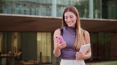 Neşeli Genç Bir Kadın Elinde Not Defteri Tutarken Akıllı Telefonuyla Nişanlı. Cep telefonuyla Mutlu İfade ve Etkileşimler sergiliyor. Genç kız internette gezinmek için cep telefonu kullanıyor.