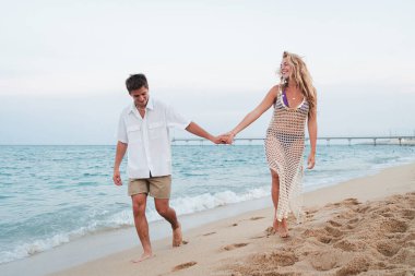 Kumsalda el ele yürüyen neşeli çift tatillerinin tadını çıkarıyorlar. Kadın, gülümseyen bir adamla birlikte okyanus dalgalarının altında parlak gökyüzünün altında eğleniyor. Yüksek kalite