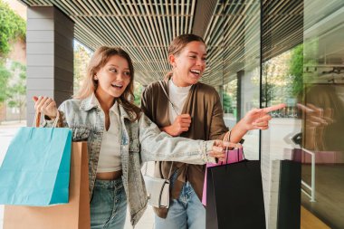 İki mutlu arkadaş birlikte alışveriş yaparak keyifli bir gün geçiriyorlar. Yeni hazineleriyle dolu renkli alışveriş torbalarını tutarken bir mağaza vitrinine heyecanlı bir şekilde gülüyorlar ve işaret ediyorlar.