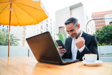 Başarılı Olgun İş Adamı Akıllı Telefon ve Bilgisayarı İş İçin Kullanırken Sevinç İfade Ediyor. Hedeflere Ulaşmak ve İş Başarısında Yeni Tepelere Ulaşmak İçin Güven ve Kararlılık Gösteriliyor. Yüksek kalite