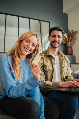 Mutlu çift, kredi kartlarını online alışveriş yaparken rahat bir ev ortamında dizüstü bilgisayarlarıyla eğleniyor, uygun alışverişleri zahmetsizce ve neşeyle yapıyorlar.