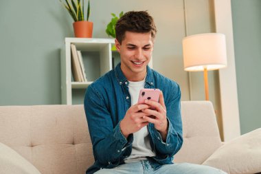 Parlak bir oturma odasıyla çevrili rahat bir koltukta dinlenirken cep telefonuyla gezen neşeli adam. Evde cep telefonu kullanarak eğlenen genç bir adam. Ergen, Smartphone 'unu izliyor. Yüksek