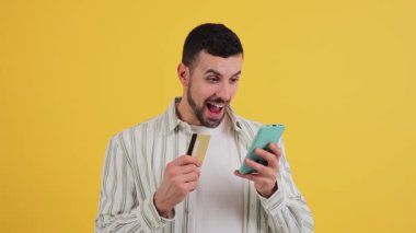 Elinde kredi kartı olan heyecanlı bir genç adam sarı arka planda online alışveriş yapmak için akıllı telefon kullanıyor. Kartı kameraya coşkuyla ve neşeli bir ifadeyle gösteriyor. Yüksek kalite 4k