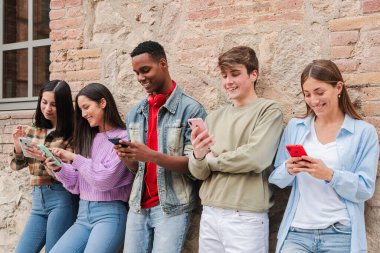 Beş hevesli genç arkadaş akıllı telefonlarına dalmış, kahkahalarını paylaşıyor ve eğlenceli mobil etkileşimlere giriyorlar, gençler arasında canlı, sosyal bir ortamda modern iletişimi sergiliyorlar.
