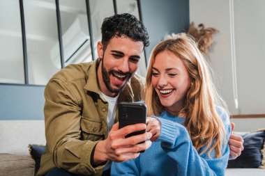 Neşeli bir çift, eğlenceli uygulamaları ve heyecan verici içerikleri keşfetmek için akıllı telefonu kullanırken aynı zamanda eğlenceli anları paylaşıyor kahkaha ve bağlantılarla dolu sıcak bir ev ortamında. Yüksek kalite