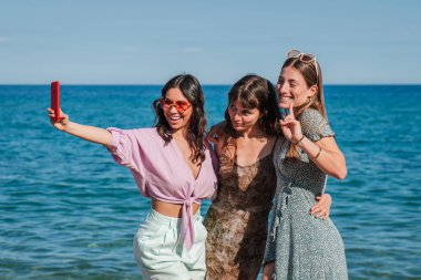 Üç neşeli genç kadın sahilde mutlu bir şekilde selfie çekerken güneşli bir günün tadını çıkarırken kahkaha ve arkadaşlıkla birlikte sosyal medya için akıllı telefonlarıyla mükemmel anı yakalıyorlar.