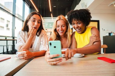 Kahve dükkanında cep telefonunu izleyen üç çok ırklı kadın. Bir grup genç kız, mutlu bir anı gülümseyen ve kaynaşan bir akıllı telefon kullanarak paylaşıyorlar. Kadınlar cep telefonuyla eğleniyor.