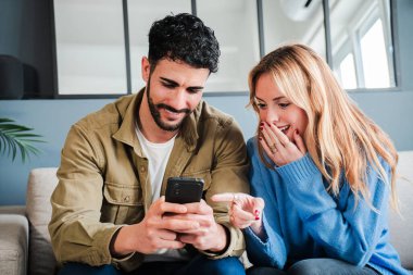 Heyecanlı çift, rahat bir kanepede otururken akıllı telefonda şaşırtıcı bir şey keşfederek sevinçle neşeli bir şekilde bağlantının iç açıcı ifadesiyle hoş bir anı paylaşıyorlar. Yüksek