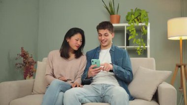 Genç çift, kanepeye rahatça otururken birlikte online alışveriş yapıyor, çeşitli uygulamaları keşfediyor ve ellerinde kredi kartlarıyla heyecan anlarını paylaşıyorlar.