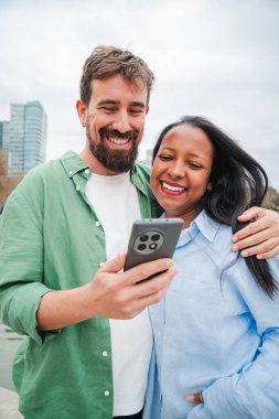 Akıllı bir telefonla meşgul olan hevesli bir çift, mobil uygulamaları keşfederken gülümseme ve kahkaha paylaşıyorlar, ilişkilerinde bağlantı ve heyecan dolu neşeli bir an yakalıyorlar.