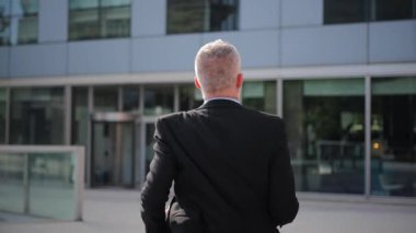 Zarif bir takım elbise giymiş kararlı bir işadamı, önemli bir ofis binasına doğru emin adımlarla yürüyor. Amacına hizmet ederken hırs ve profesyonellik sergiliyor.