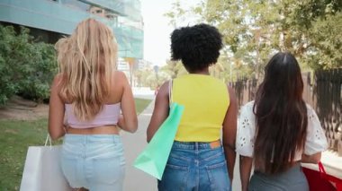 Bir grup neşeli kadın renkli alışveriş torbalarıyla birlikte yürüyorlar, gülümsemek ve kamerayla meşgul olmak için geri dönüyorlar, alışverişleri sırasında neşeli bir arkadaşlık ve keyif anı yakalıyorlar.