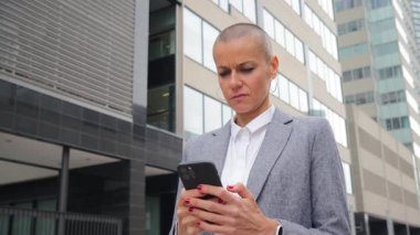 Kafası tıraşlı kararlı bir kadın akıllı telefonuyla meşgul oluyor, yoğun konsantrasyonunu ve duygusal tepkisini sergiliyor.