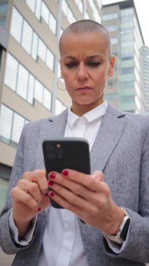 Kafası tıraşlı kararlı bir kadın akıllı telefonuyla meşgul oluyor, yoğun konsantrasyonunu ve duygusal tepkisini sergiliyor.