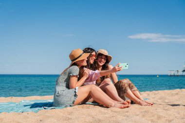 Sahilde akıllı telefonlarıyla selfie çekerken hatırlanmaya değer bir deneyimi paylaşan neşeli üç kadın arkadaşlıklarını ve birlikte geçirdikleri yaz anlarının keyfini sergiliyorlar. Yüksek